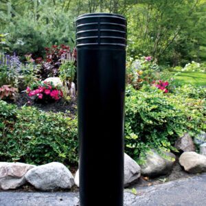 A Vestil Cinco-Black Bollard Cover 52 In stands on a paved surface with stone-bordered landscaping. Lush greenery and colorful flowers are abundant in the background under a canopy of trees.