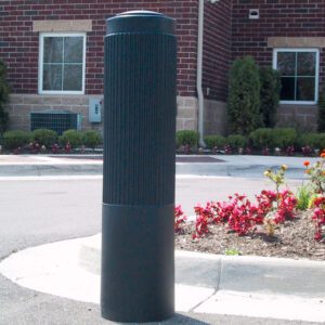A tall, cylindrical black bollard with a Vestil Rib-Black Bollard Cover 52 In stands on concrete by the curb, surrounded by red flowers and greenery. A brick building with windows is visible in the background.
