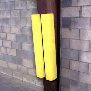 A steel pole, guarded by two yellow Vestil Polyethylene Ultra-I-Beam Protectors, stands in a corner against a brick wall and smooth concrete floor, ensuring safety and durability.