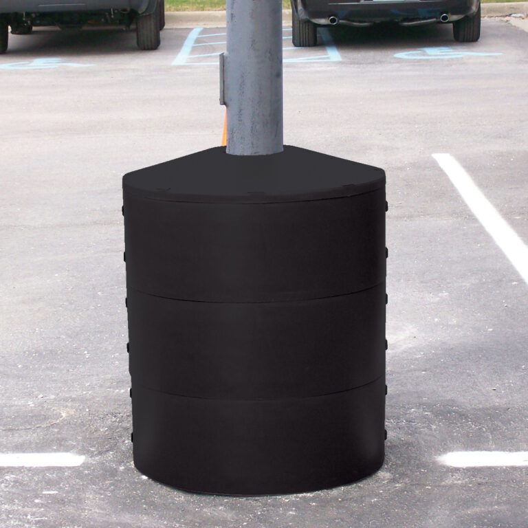 LPBP-24-BK_A.jpg A Vestil Light Pole Base Protector Black surrounds the gray lamppost base in a parking lot, forming a cylindrical barrier from three stacked segments, with parking spaces and partial car views in the background.