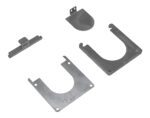 On a flat surface are four metal parts: a notched rectangular bar, a rounded cap with tab, and two U-shaped plates with corner holes, designed as additional elements for the Vestil Removable Bollard Base Additional system.
