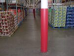 A warehouse aisle with shelves of packaged goods is centered by a Vestil Red Round Column Wrap 8 In, extending from floor to ceiling. The concrete floor stretches to the end of the aisle, enhancing visibility.