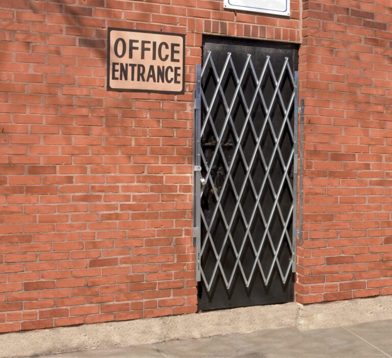 A brick wall features an Office Entrance sign above a black door, safeguarded by a Vestil Galvanized Scissor Gate-Expanded 57 In. The concrete ground and light mortar outlining the bricks enhance the sturdy, utilitarian charm.