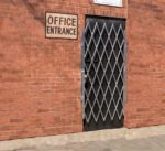 A brick wall with a sign reading Office Entrance above a Vestil Galvanized Scissor Gate-Expanded 75 In, featuring a diamond pattern. The gate stands out against the buildings red brick surface.
