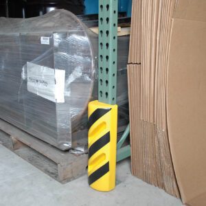 A Vestil Poly Corner Protector 15-1/2 In Height, a yellow and black striped safety guard, wraps around the base of a warehouses metal shelving unit. Nearby lie stacks of wrapped pallets and large sheets of cardboard.