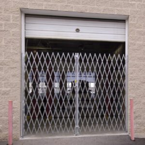 A Vestil Double Fold Scissor Gate Galv 120L X 84H secures a garage door on a beige brick building, flanked by red bollards, with industrial equipment visible inside.