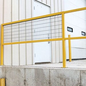 A Vestil Steel Sq Safe Handrail Wire Mesh 96 In stands in front of a white industrial building. Positioned on a concrete platform, it reaches a closed white door with a small step, while two yellow bollards nearby enhance the secure setup.