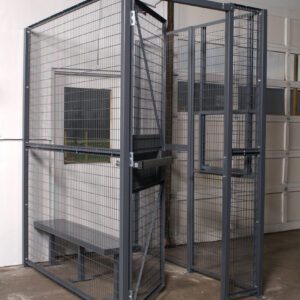 A 3-sided Vestil Driver/Trucker Access Cage 4X8 features a metal wire mesh security enclosure with a door, housing a small bench on the concrete floor. Its positioned next to a white wall with a window while a roll-up door is visible in the background.