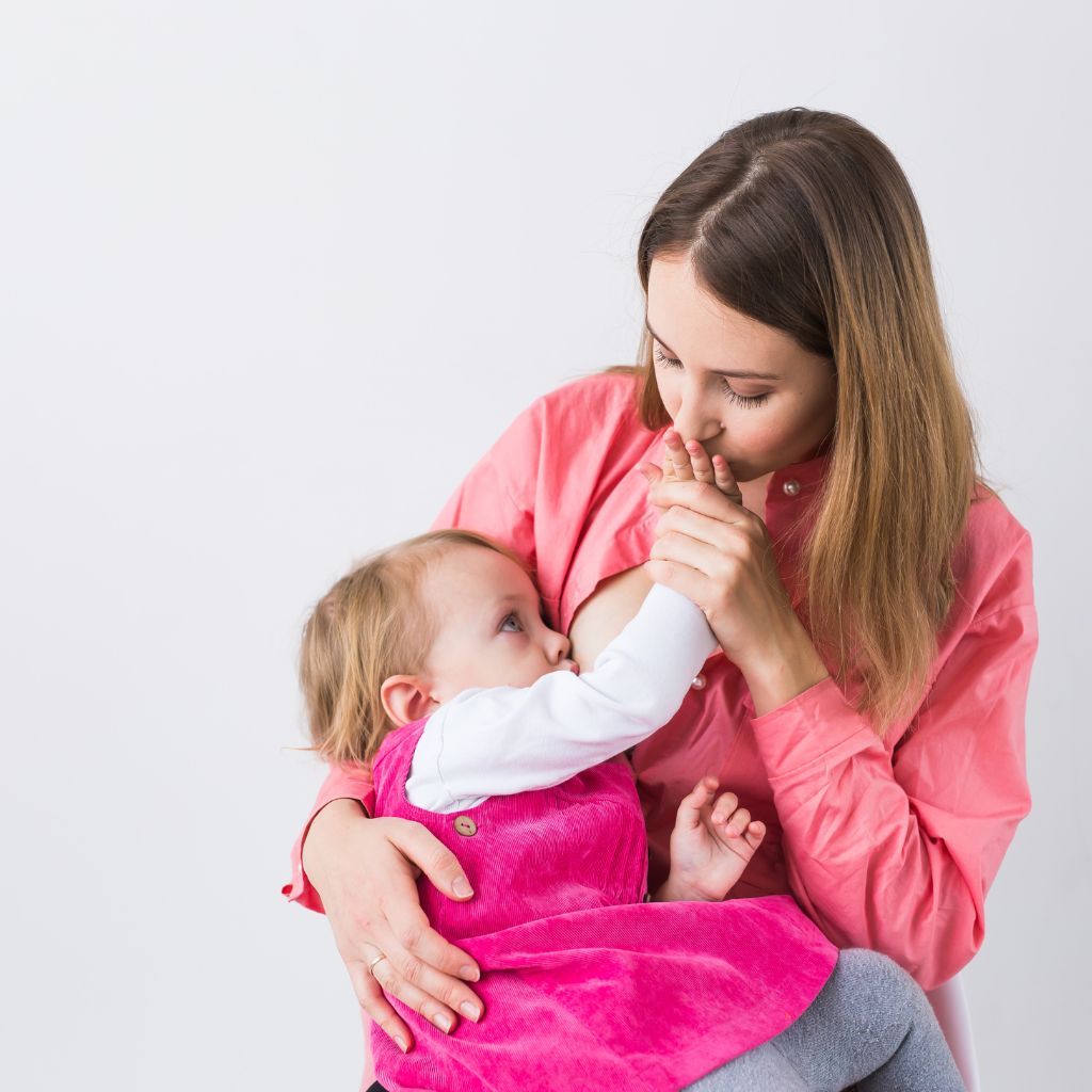 A mother in a pink shirt is using an ADA Lactation Pod while breastfeeding her baby, who wears a pink outfit with a white shirt. Sitting against a plain white background, they share a tender moment, benefitting from modern comforts designed for privacy and convenience.