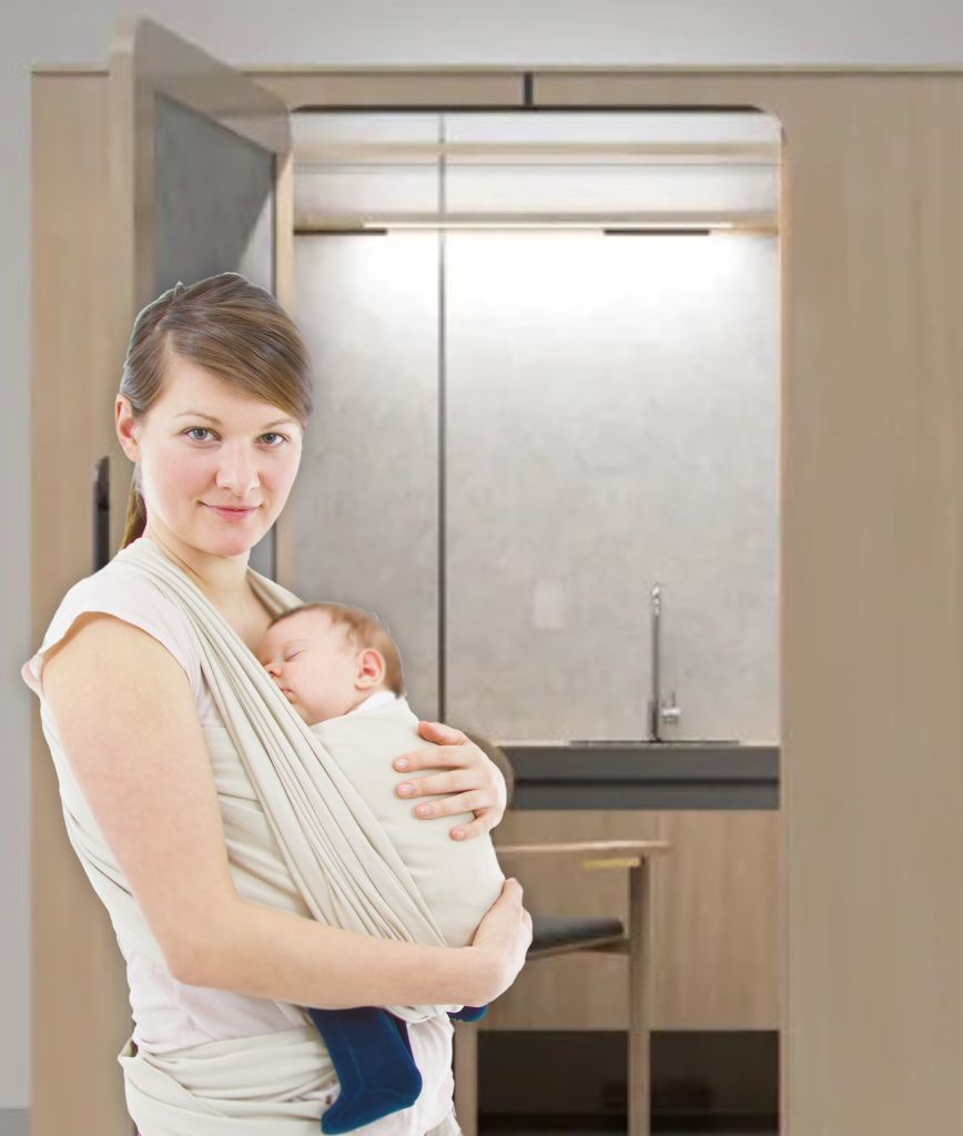 A woman stands in a modern room with a sink and cabinet, carrying a sleeping baby in a pale sling. Inspired by the PUMP Act, she appreciates the convenience of breastfeeding pods in airports, which align with the rooms soothing light color palette.