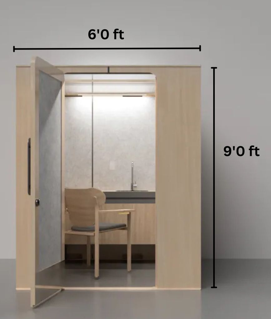 A small, light wood 6x9 ft office pod with an open door reveals a simple interior. It includes a chair facing a desk with a lamp under a shelf and features ADA-compliant elements like a back mirror extending to the ceiling. Ideal for lactation pod service plans, its set against a plain gray wall.