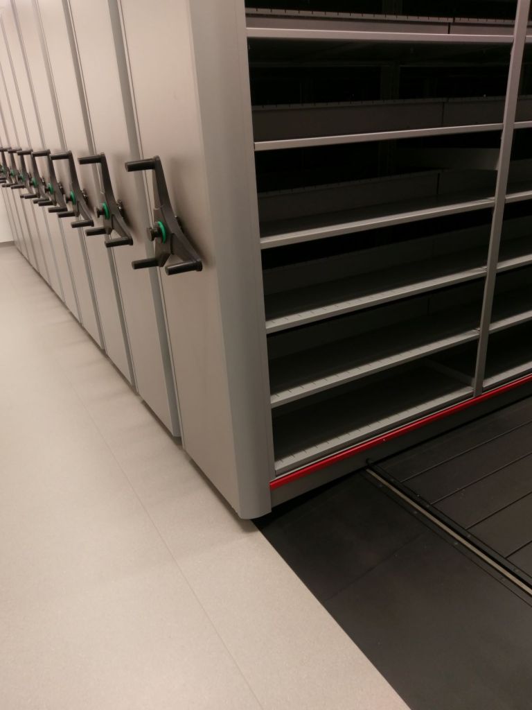 Movable metal shelving units with crank handles in a modern storage facility offer efficient medical records organization. The mostly empty units reveal shelves and a track system on the floor, designed for seamless health information management storage.