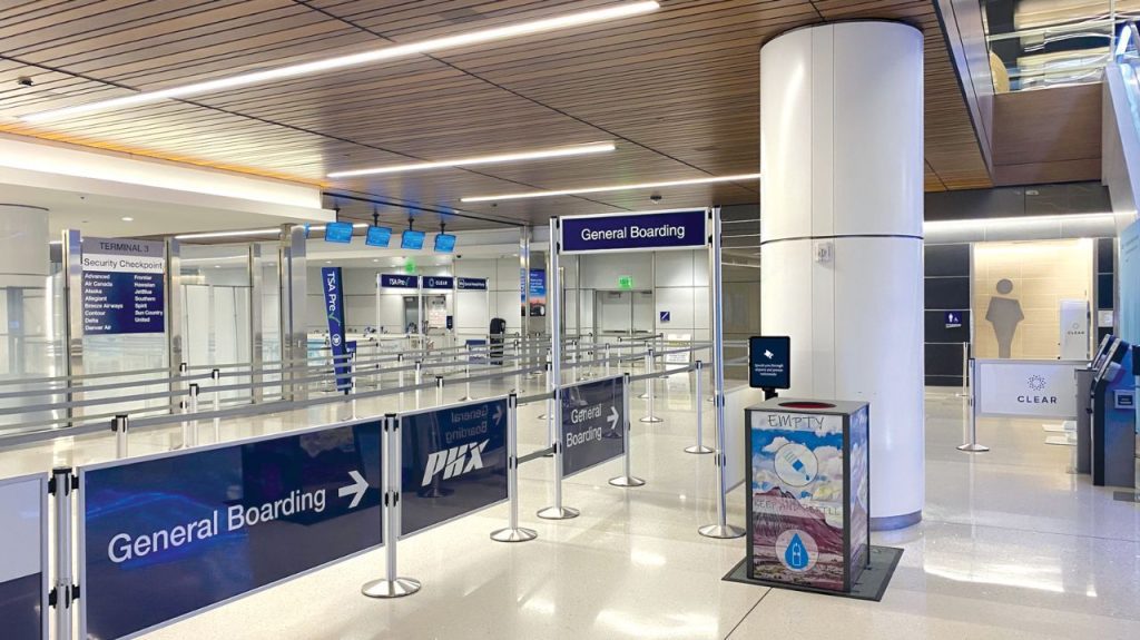An empty airport security checkpoint area with signs for general boarding, stanchions, digital screens, and a recycling bin. The area is brightly lit with a wood-paneled ceiling and tiled floors.