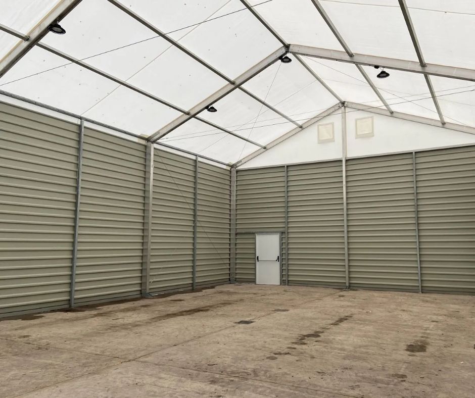 A large, empty modular warehouse design with beige metal walls, a translucent roof, and a single closed door centered on the far wall. The concrete floor and bright lighting make it ideal for rapid deployment warehouse needs.