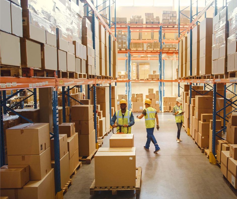 A spacious warehouse with tall shelves stacked with boxes, where workers in safety vests and helmets use a pallet jack along a wide aisle—demonstrating flexible warehouse design for efficient multi-client warehouse solutions.
